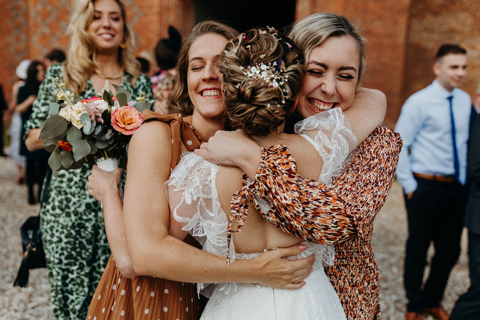 Natural wedding moment of Bride hugging two cheerful friends with bouquets, surrounded by guests in the background.