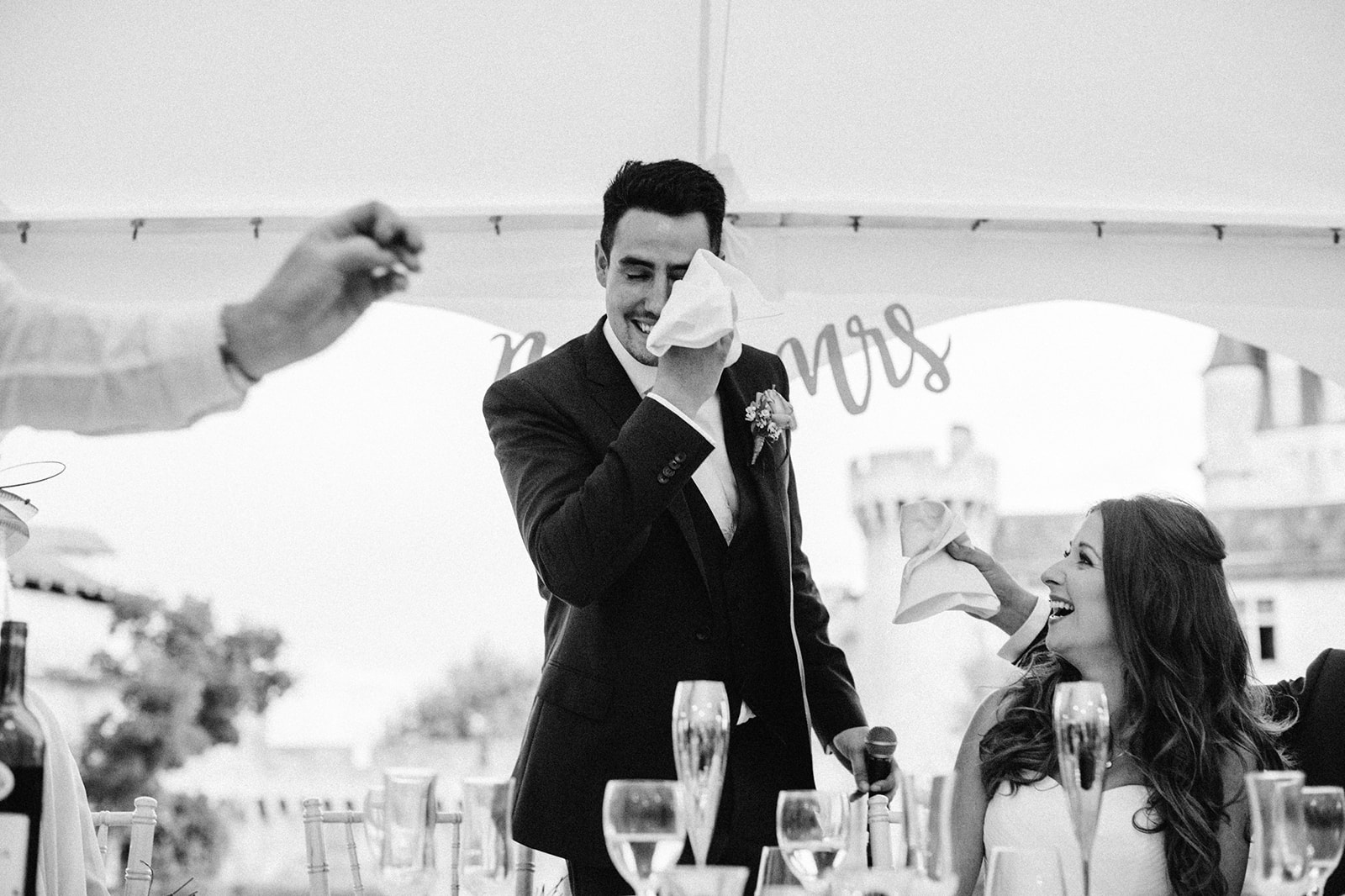 Award-winning candid photograph of a groom wiping tears with napkins passed by guests as bride laughs by his side.
