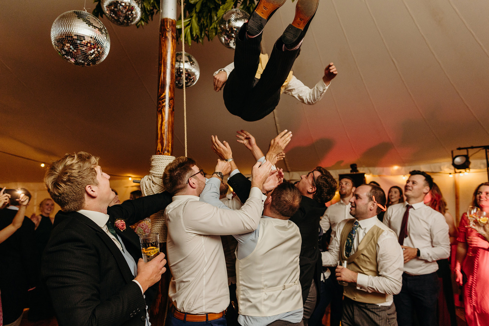 Exuberant wedding reception moment: Man tossed in the air by cheering guests on the dance floor inside a lively tent.