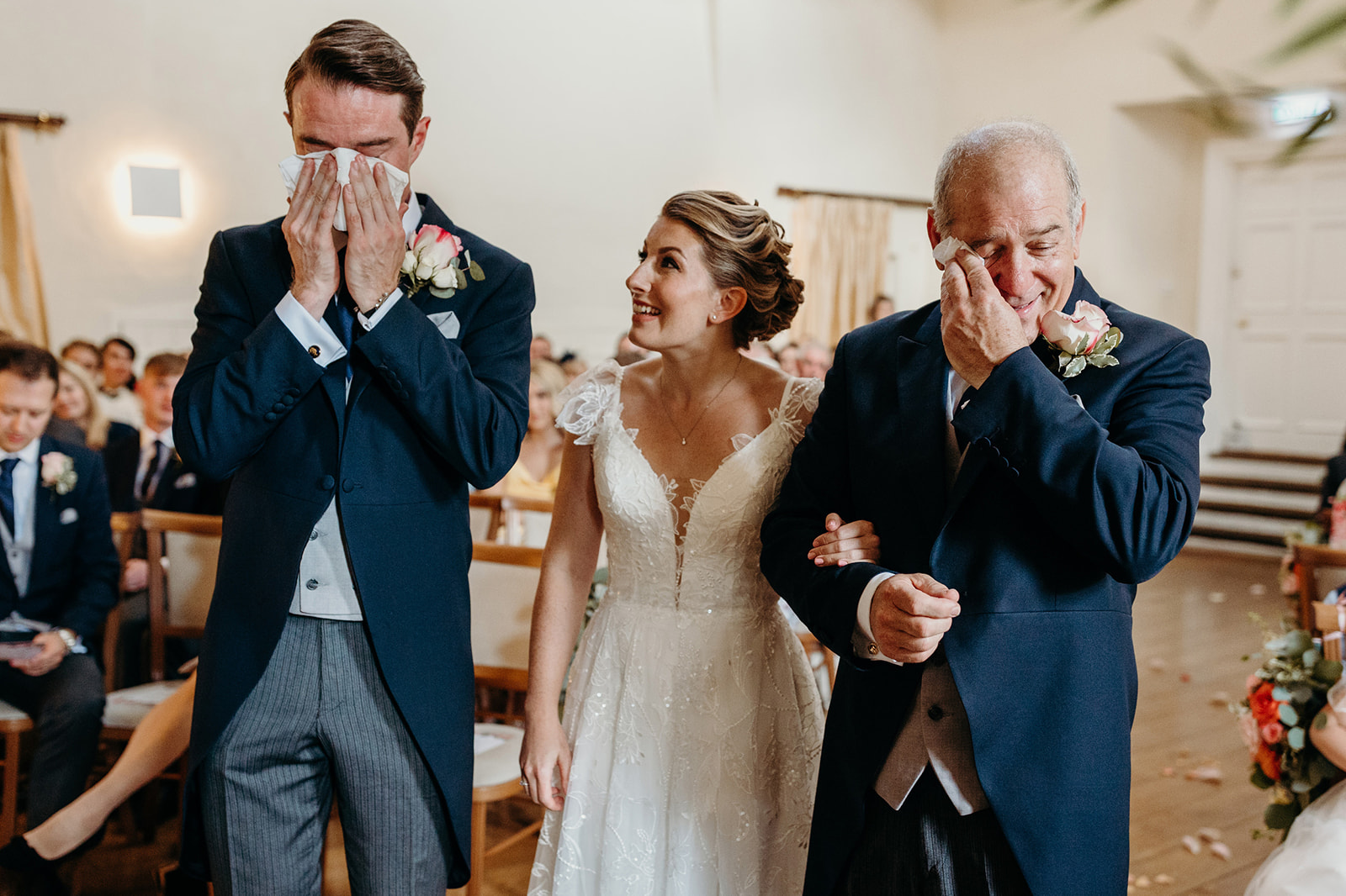 Documentary wedding photograph of a bride walking down the aisle with her smiling father, as the groom covers his face, 