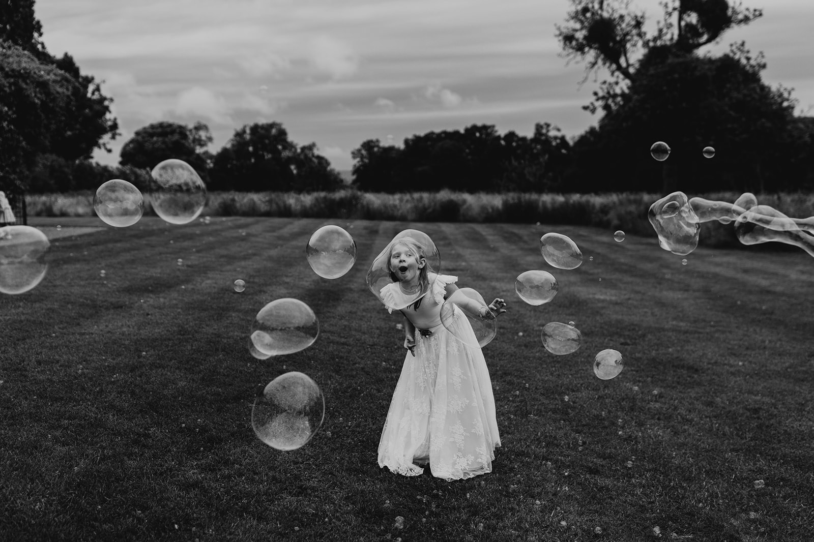 Photojournalistic wedding image depicting a young girl in a white dress enjoying large soap bubbles in a field