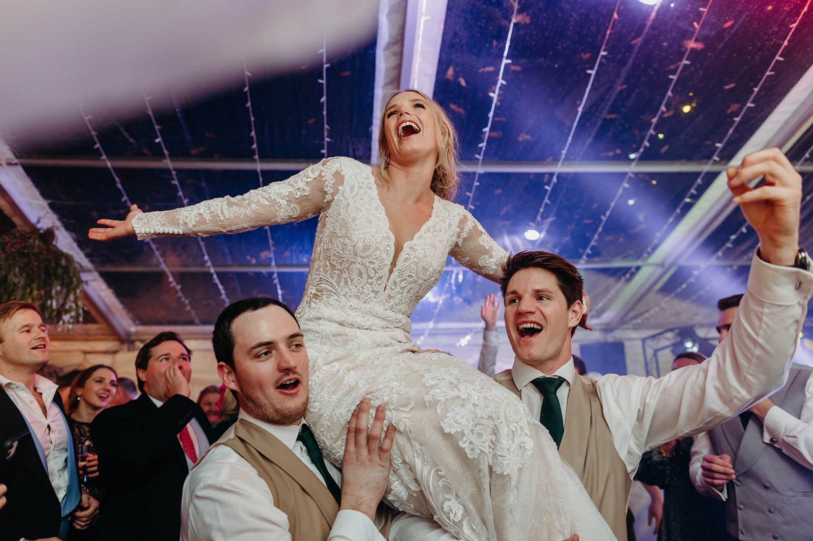 Lively celebration as bride is hoisted by groom and guest at a festive wedding reception beneath a string-lit tent.