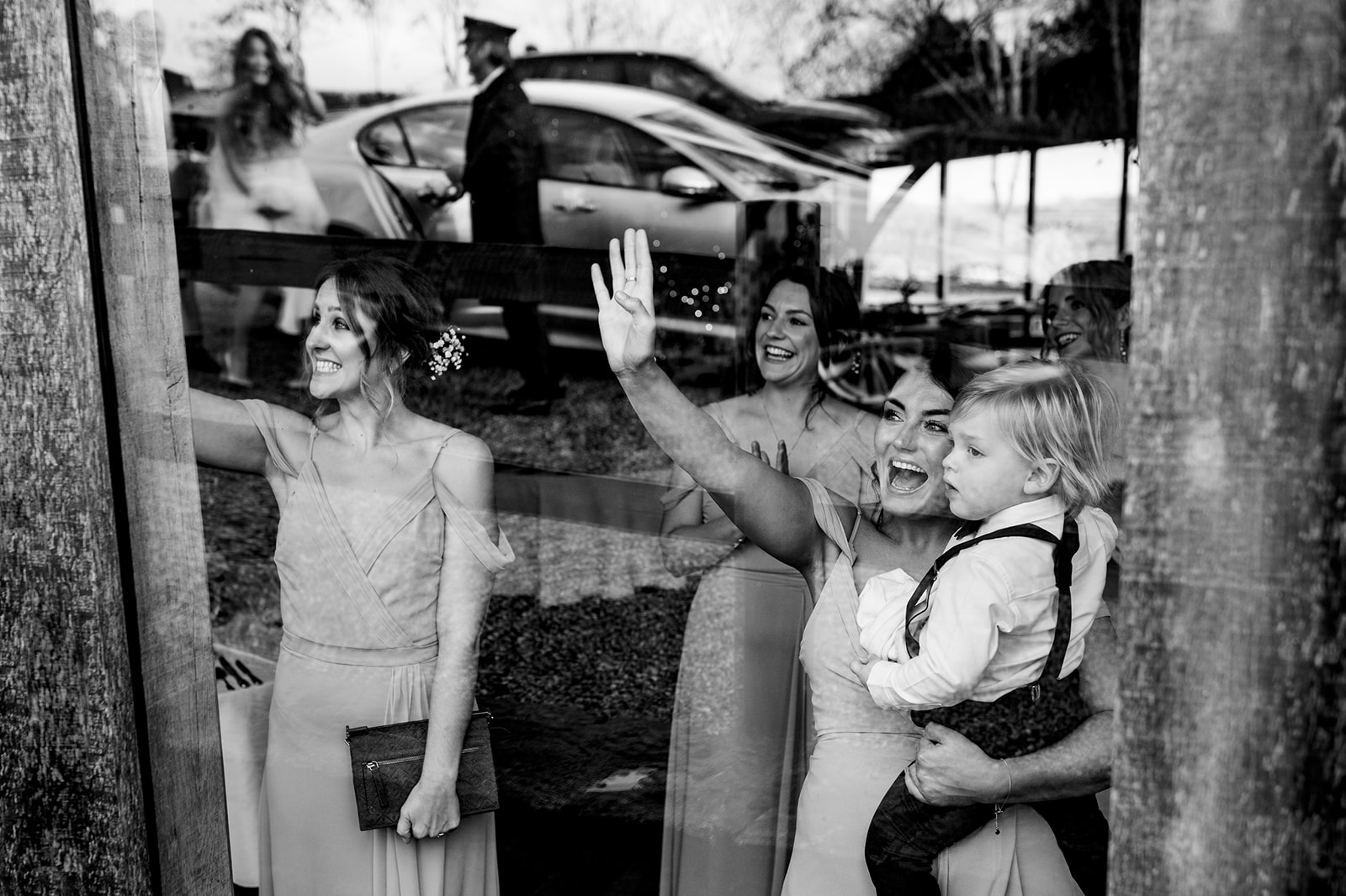 Candid black and white photograph of bridesmaids and a child waving to the bride, captured in the venue window's 