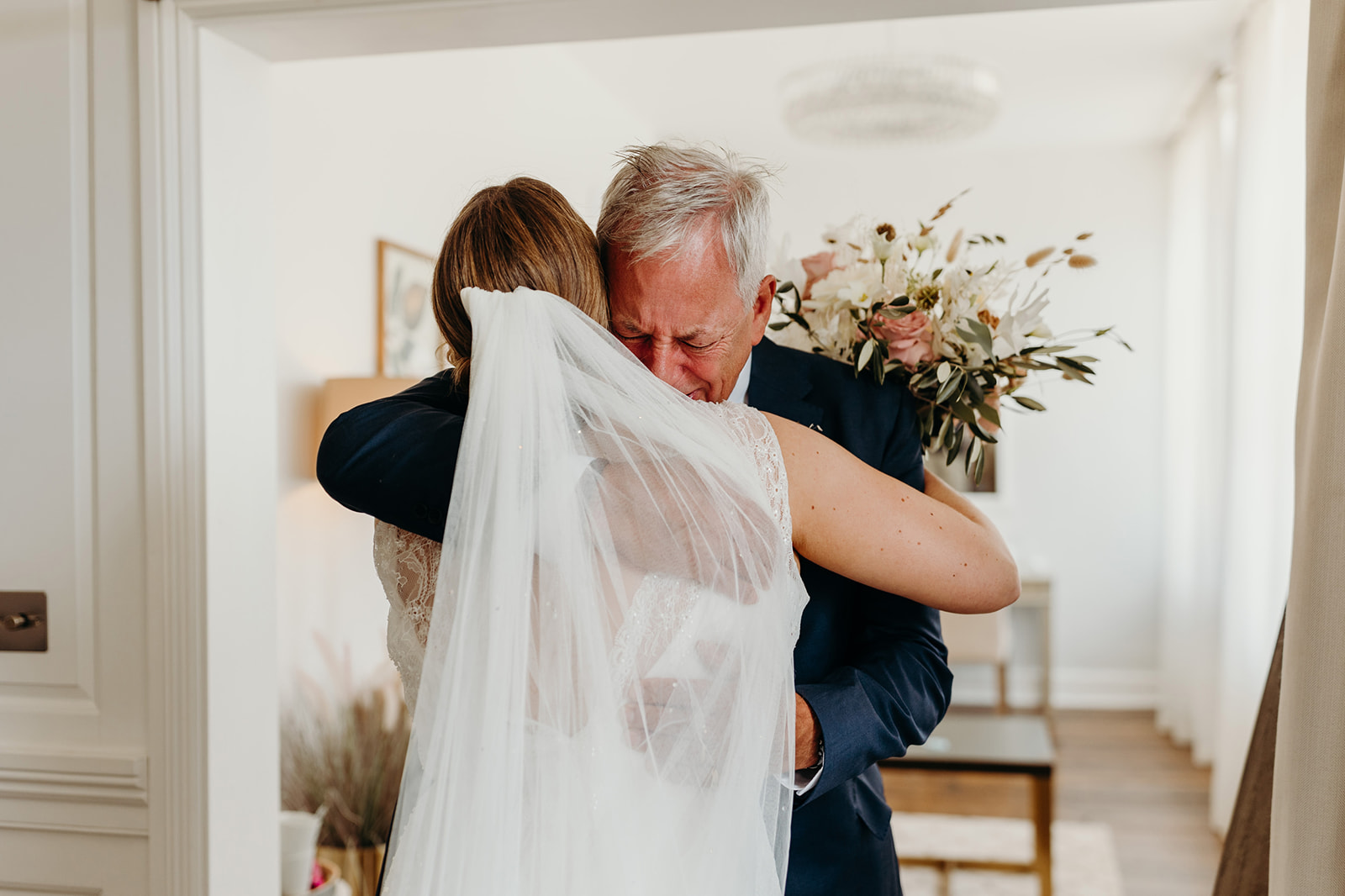 Tender embrace captured as a father hugs his bride daughter in a warmly lit room, both emotional as he fights back tears
