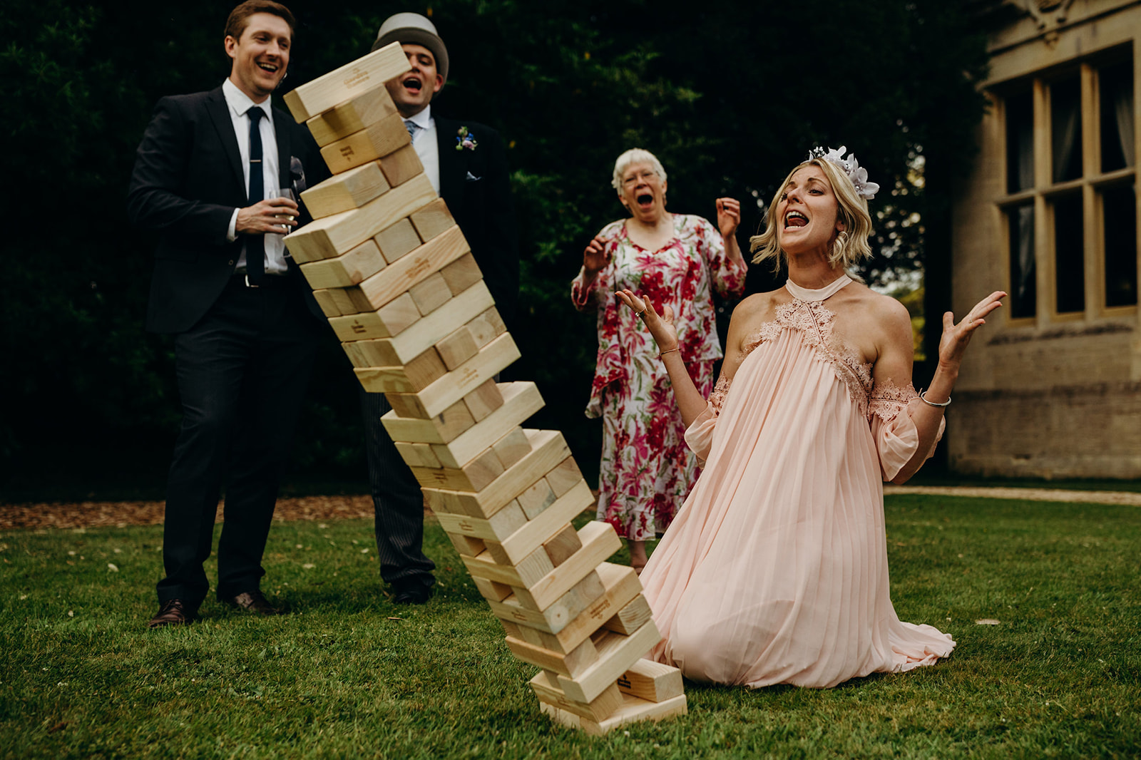 Woman in pink laughs as giant Jenga tower falls during drinks reception, guests laugh in background 