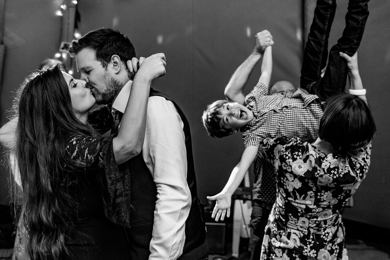 Natural black and white photograph of a couple kissing at a wedding party, as children play and an adult lifts a child 