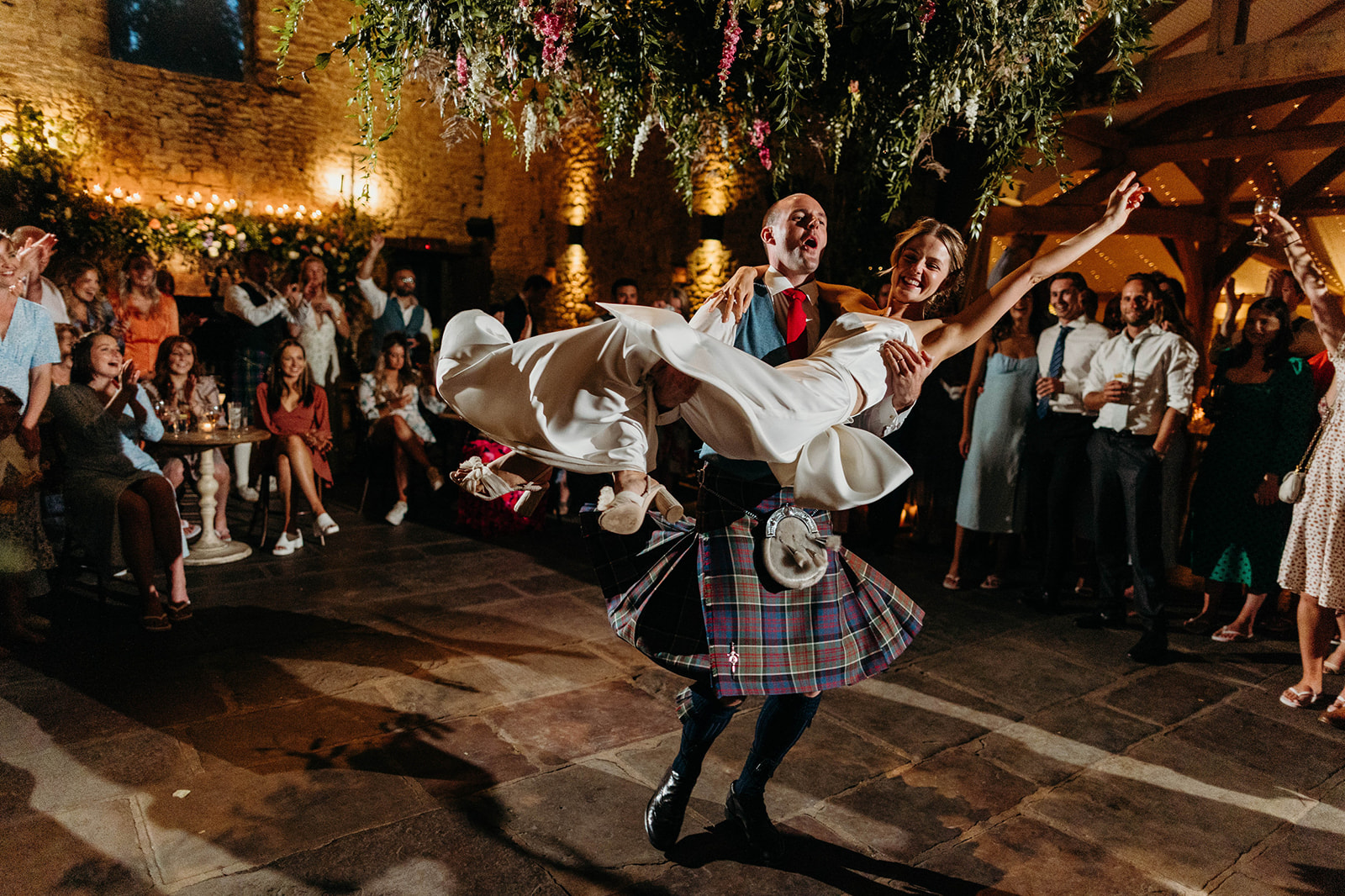 Documentary style image depicts a kilt-wearing groom carrying his bride across the dance floor, with guests applauding