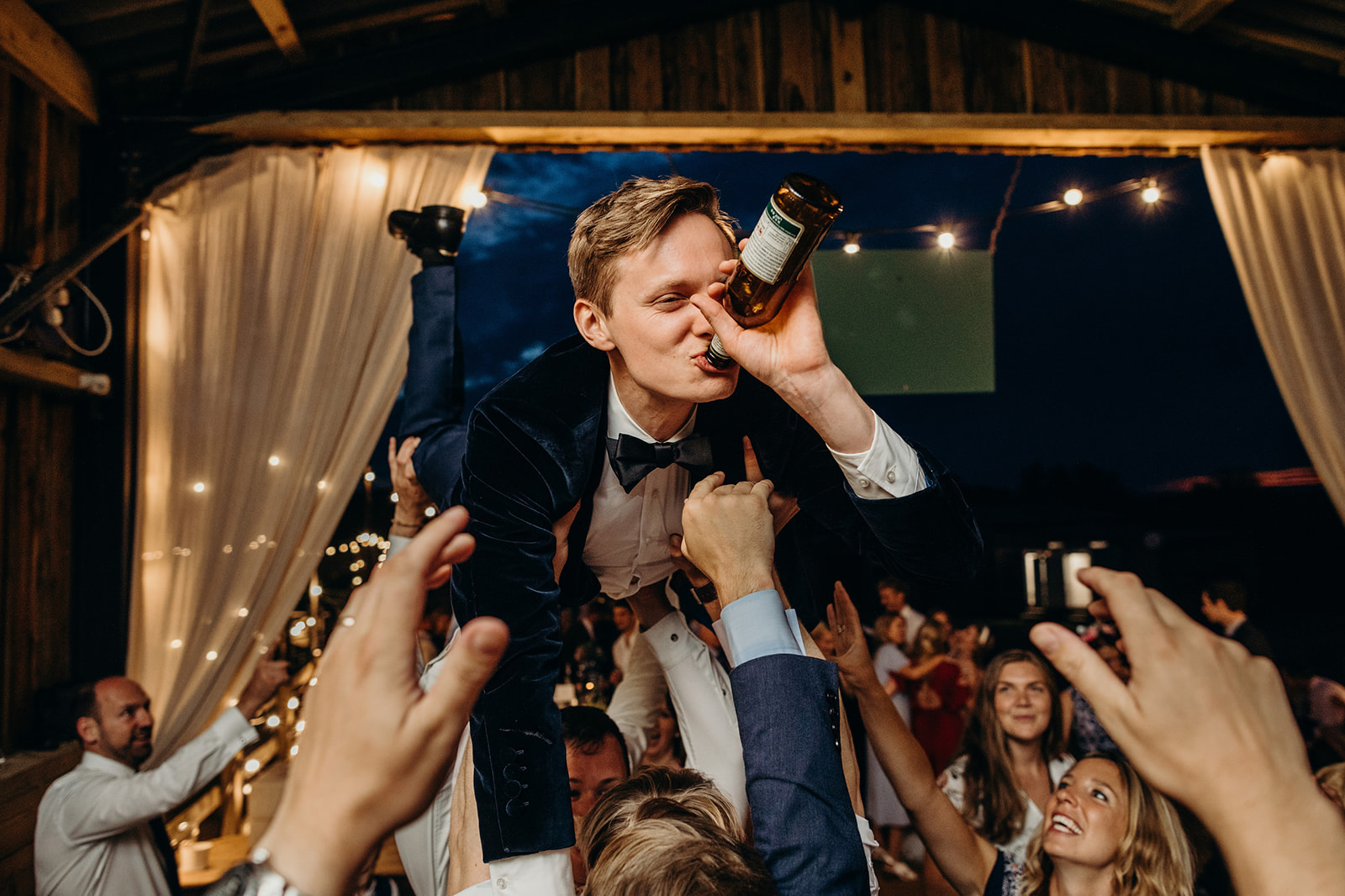 Documentary style wedding image depicts a groom, held aloft by guests, drinking from a bottle as onlookers applaud 