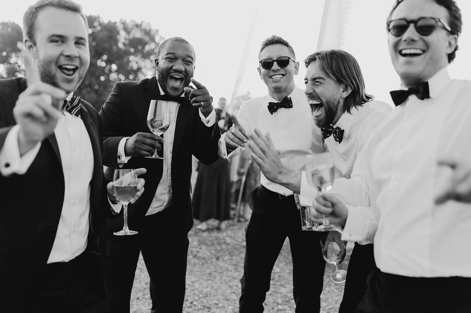 Cheerful moment captured: Four tuxedo-clad men laugh and raise champagne glasses at a festive outdoor event.'