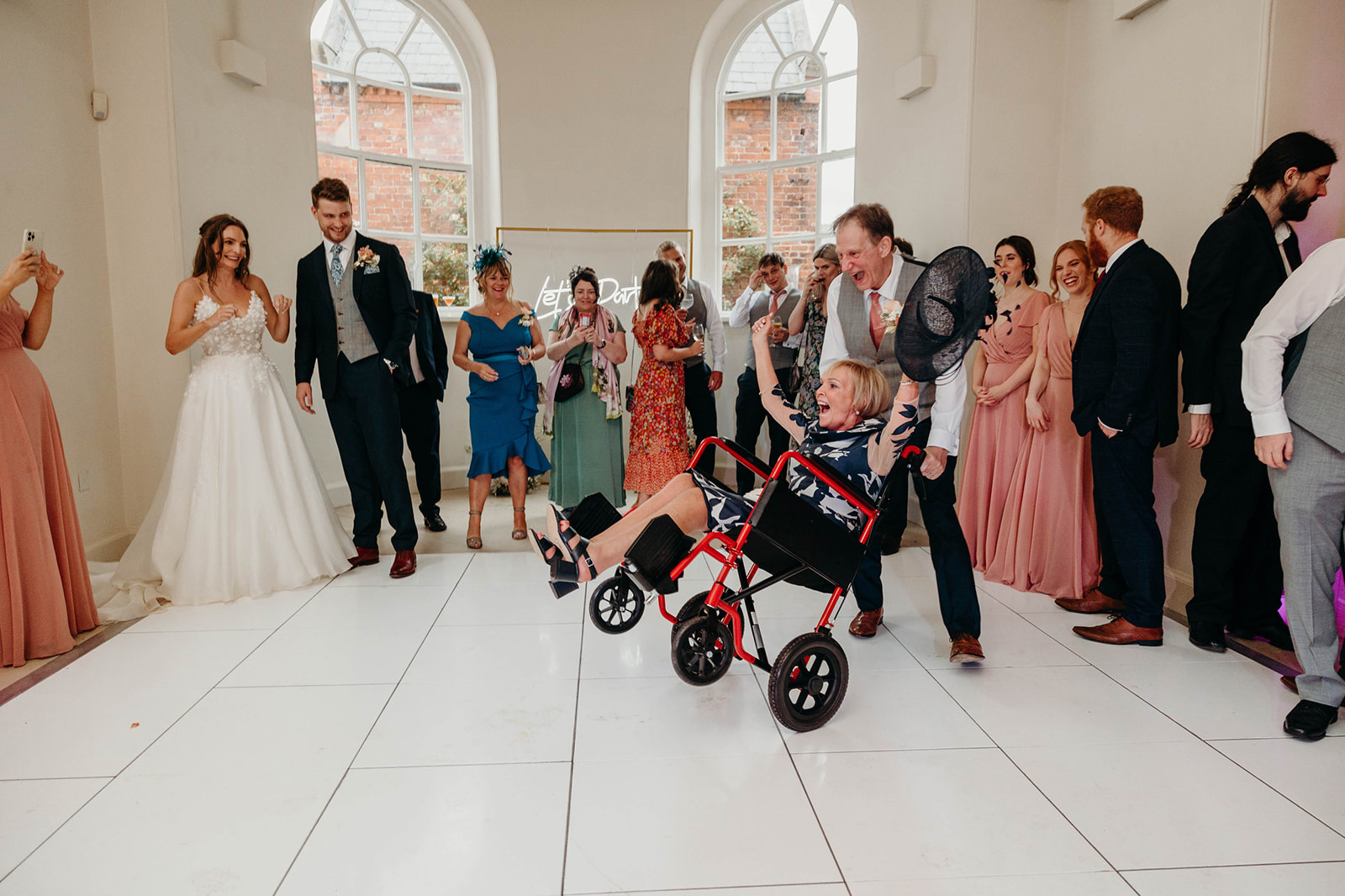 Candid shot of the groom's mother, grinning in a wheelchair, pushed enthusiastically by a man at wedding party