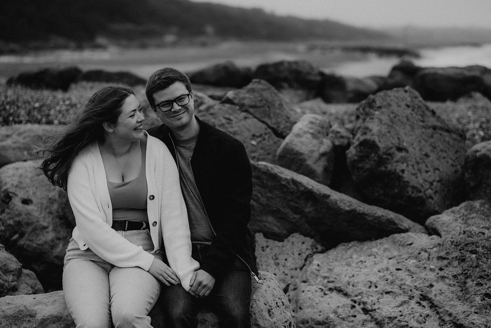 Black and white image of a couple sat on the rocks at Highcliffe Beach. Pre-wedding photoshoot. 