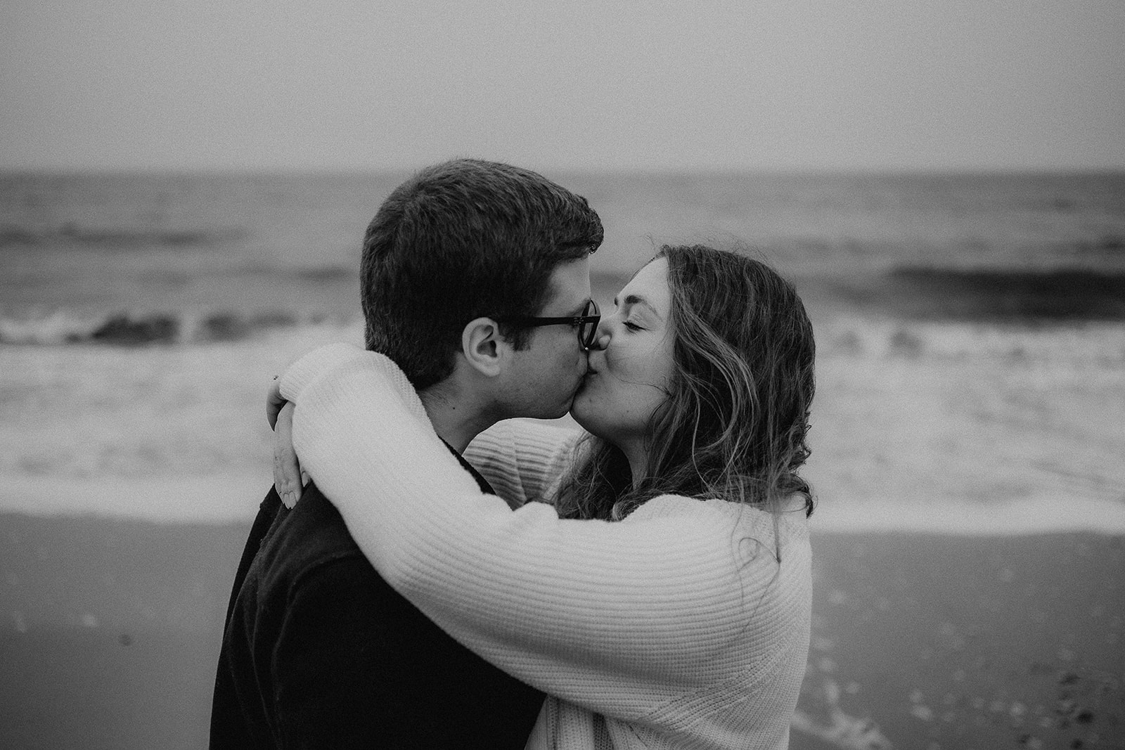 Black and white image of an engaged couple kissing on the beach in front of the waves. 