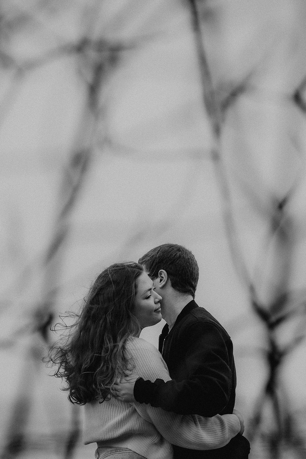 Black and white photo of a couple embracing between spidery tree branches at Highcliffe Beach photoshoot