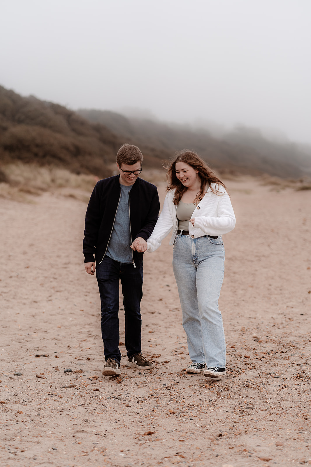 Couple laugh together as they walk hand in hand along Highcliffe Beach for their engagement photoshoot