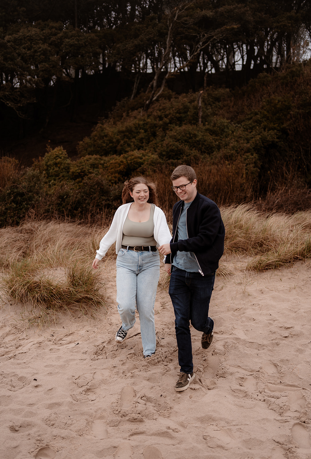 Couple run down the sand dunes hand in hand at their pre-wedding photoshoot on Highcliffe Beach