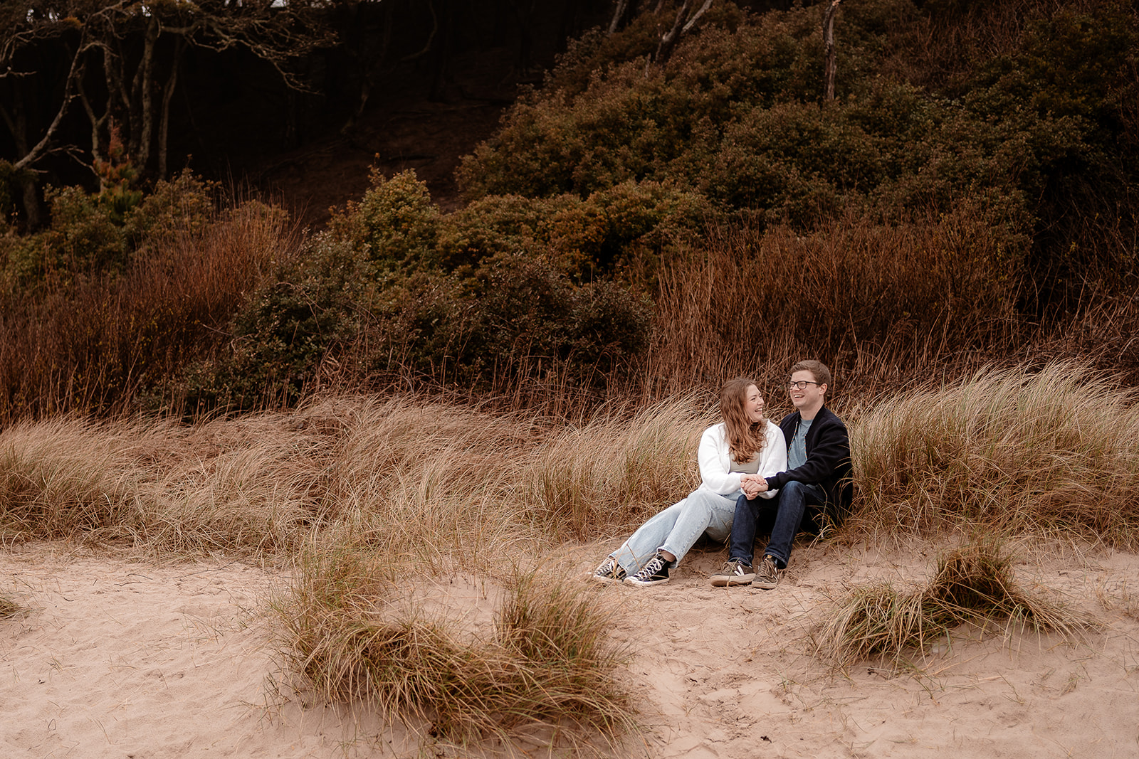 Couple sit on the sand dunes and laugh together on Highcliffe beach. Pre-wedding photoshoot. 