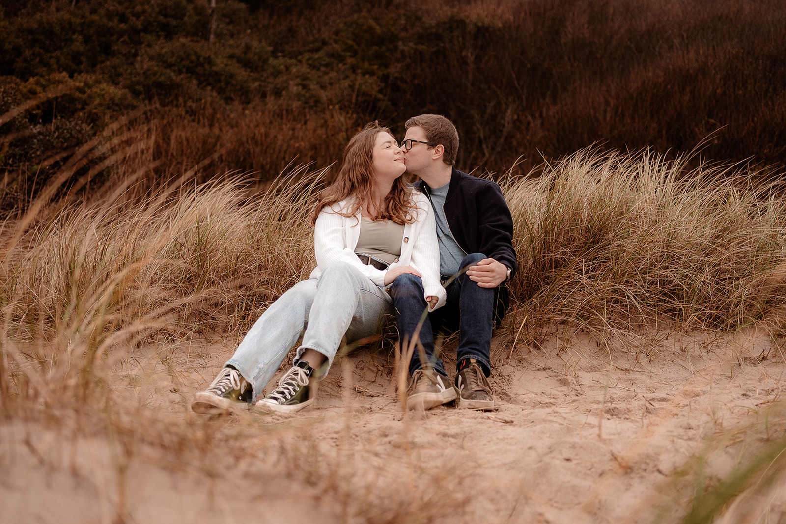 A couple sit together on the sand dunes at Highcliffe Beach at their engagement photoshoot. 