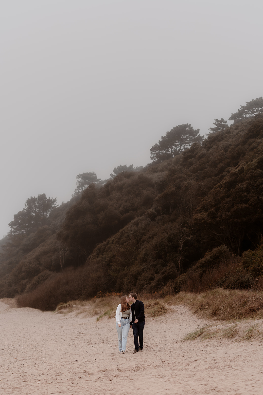 Couple walk hand in hand in front of a misty woodland and beach backdrop. 