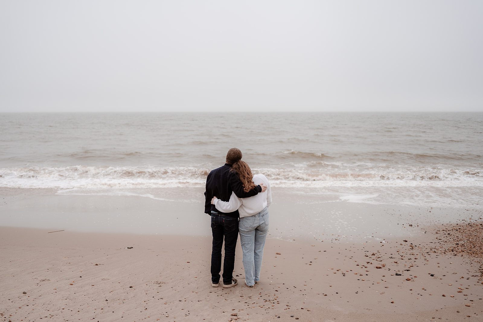 Engaged couple cuddle together and look out to sea at their Highcliffe Beach engagement shoot. 