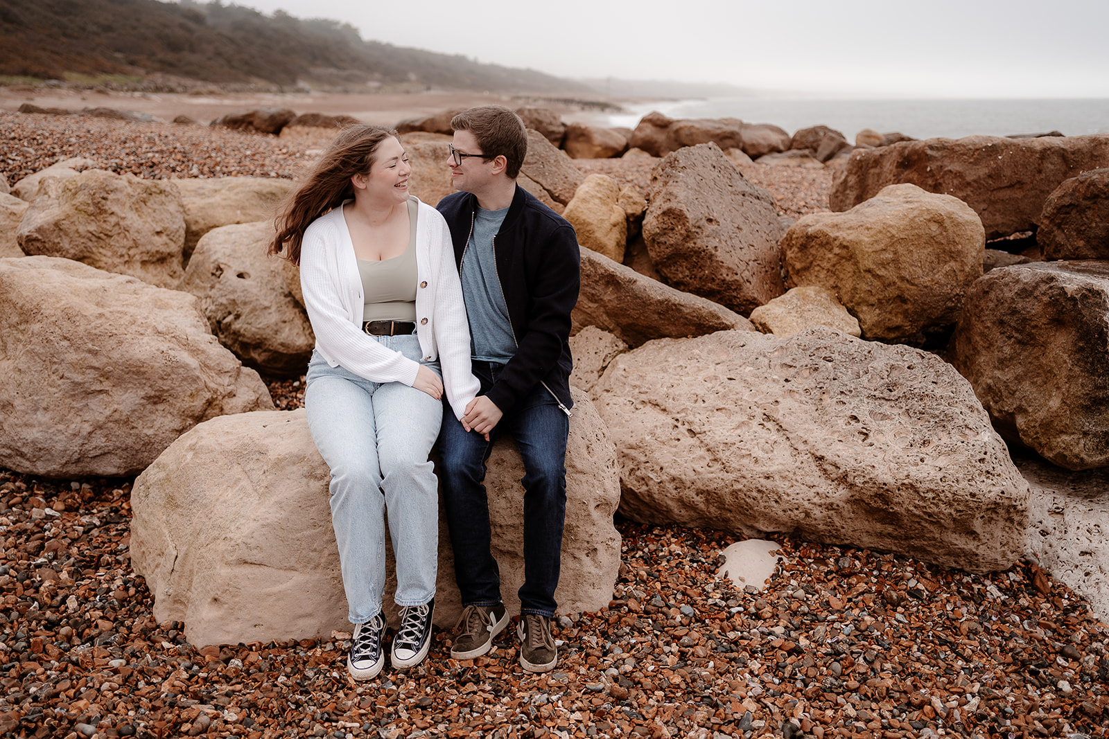 Engaged couple sit on giant rocks on Highcliffe Beach for their engagement photoshoot. 