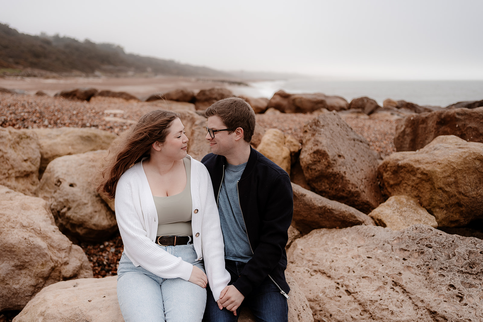 Engaged couple sit on giant rocks on Highcliffe Beach for their pre-wedding photoshoot. 