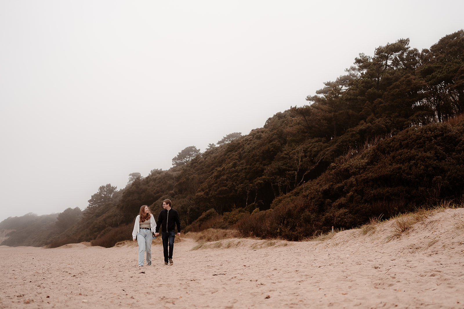 Engaged couple walk hand in hand along Highcliffe Beach at their moody engagement photoshoot. 