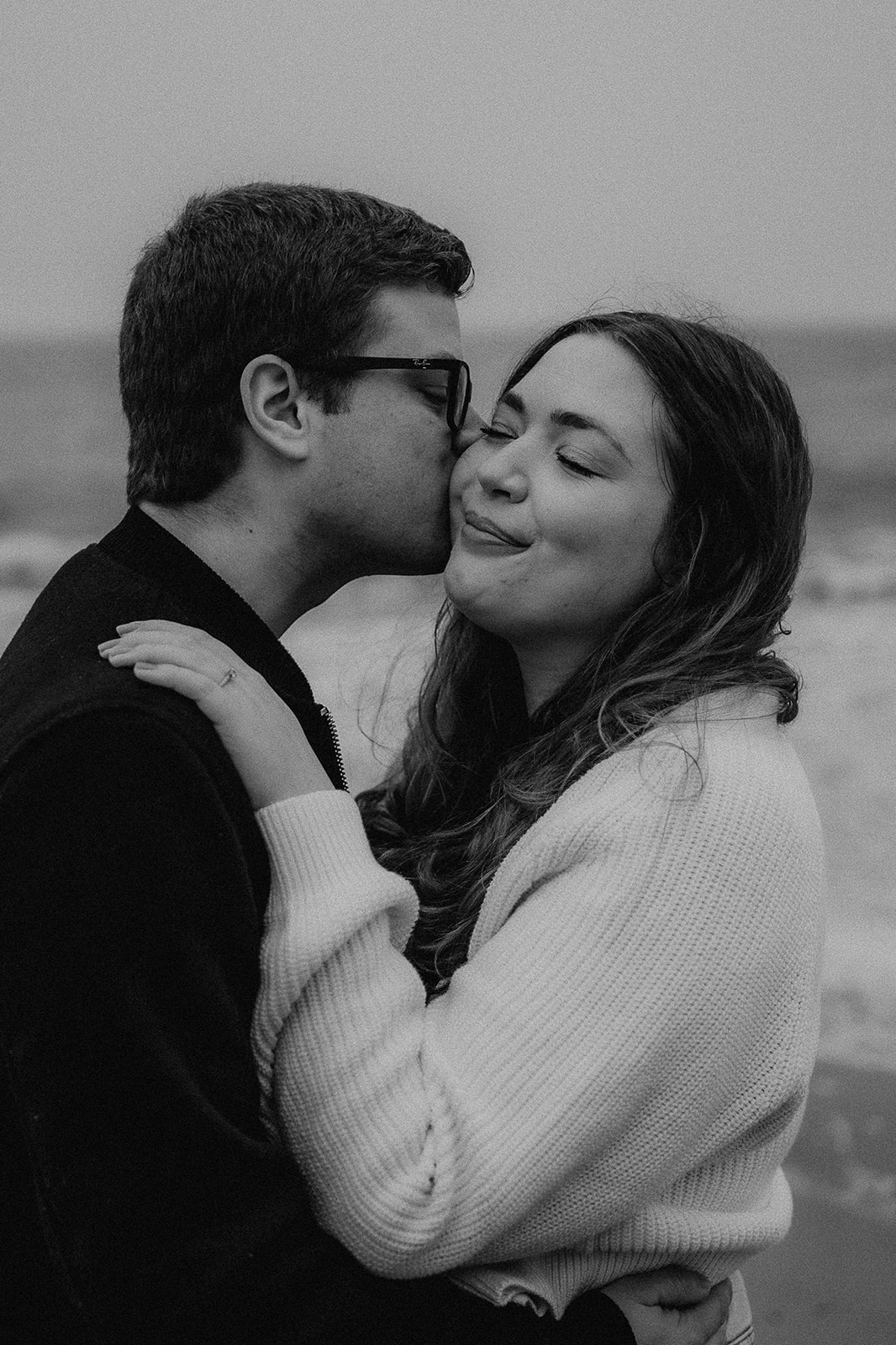A husband-to-be kisses his fiance on the cheek at their engagement photoshoot on Highcliffe Beach. 