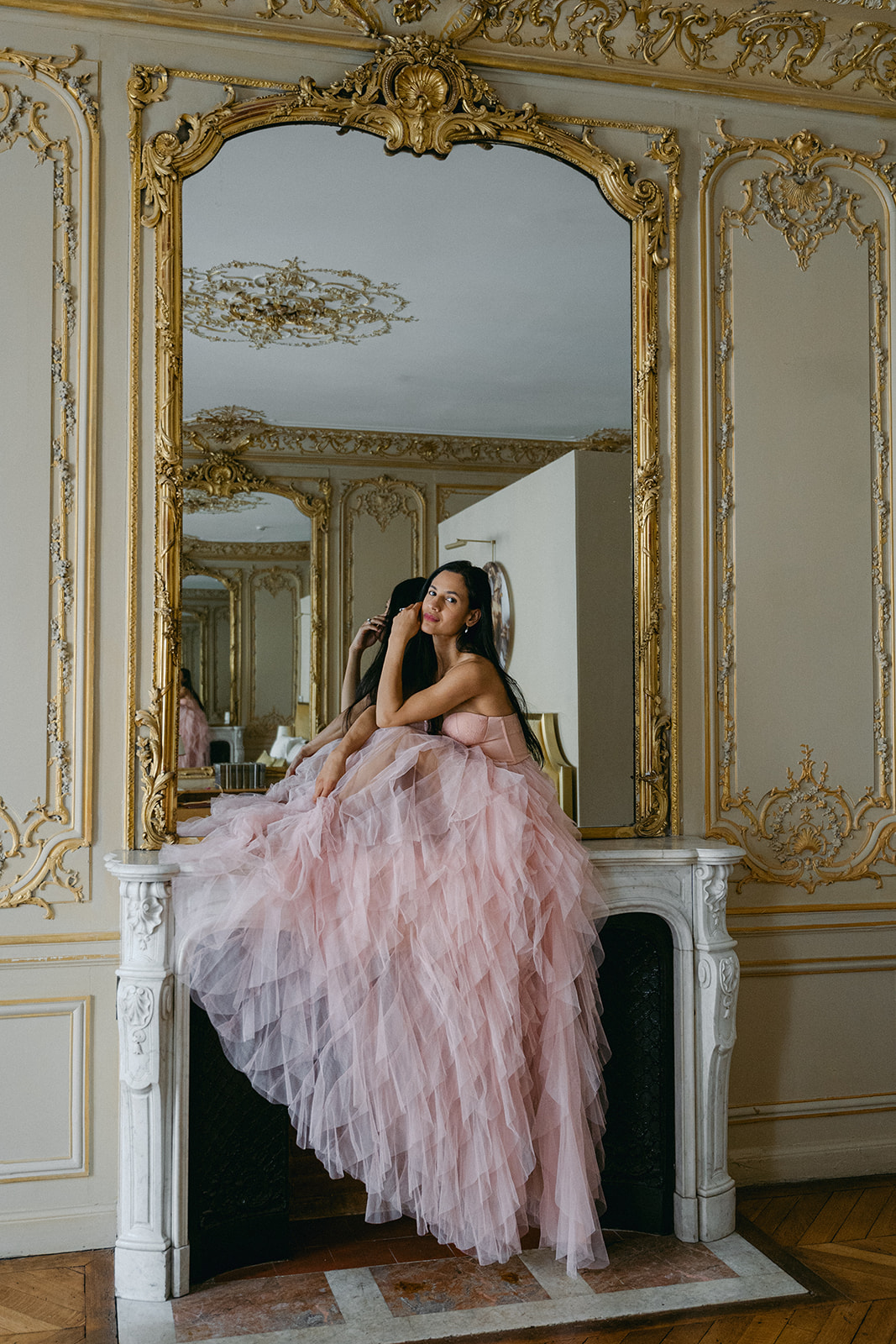 Woman in flowing pink tulle dress sits on a fireplace in Parisian hotel